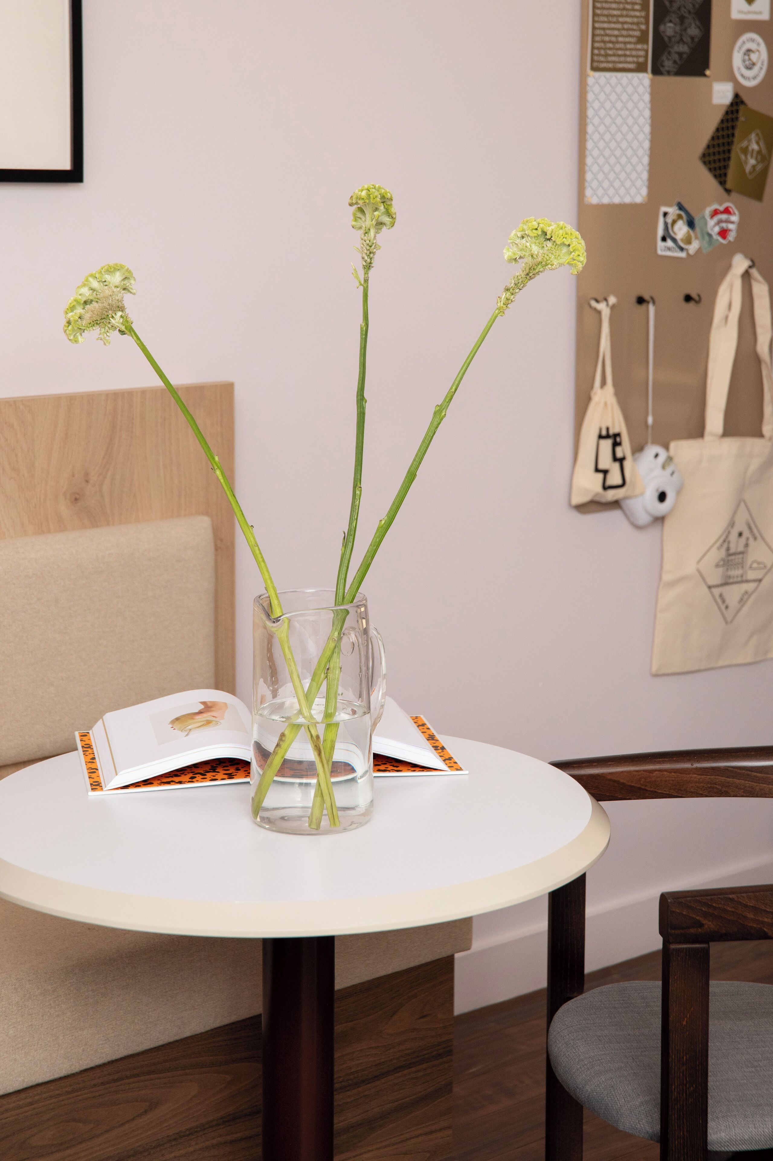 Een ronde tafel met een glazen vaas gevuld met groene stelen en kleine bloemen, een wit boek en een beige tas op tafel.