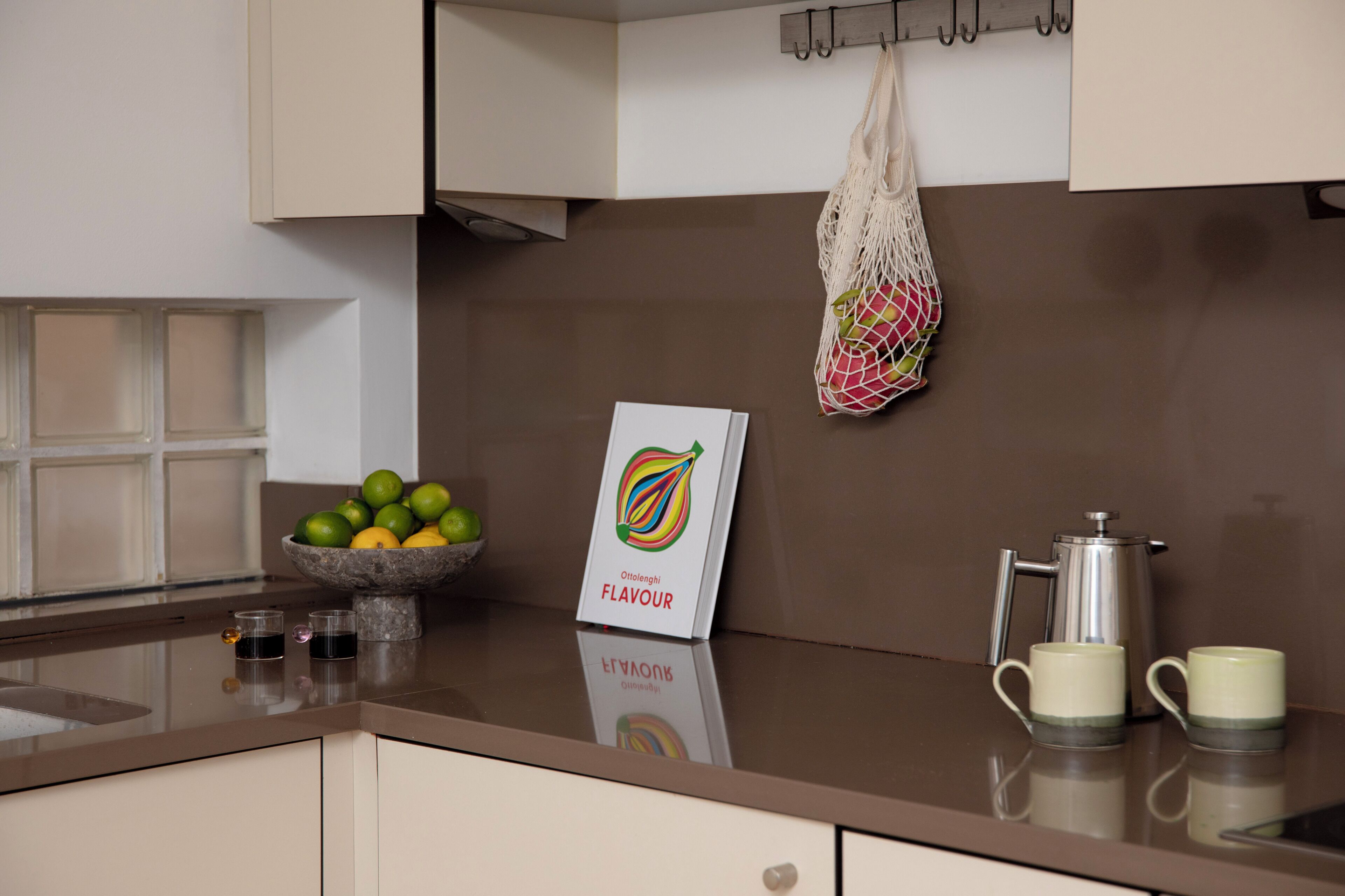 A kitchen counter with a coffee maker, two cups, a bowl of fruit, and a hanging bag.
