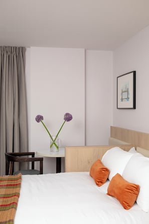Habitación de hotel con una cama, dos almohadas naranjas, una mesita con un jarrón de flores moradas, una silla y un cuadro en la pared.