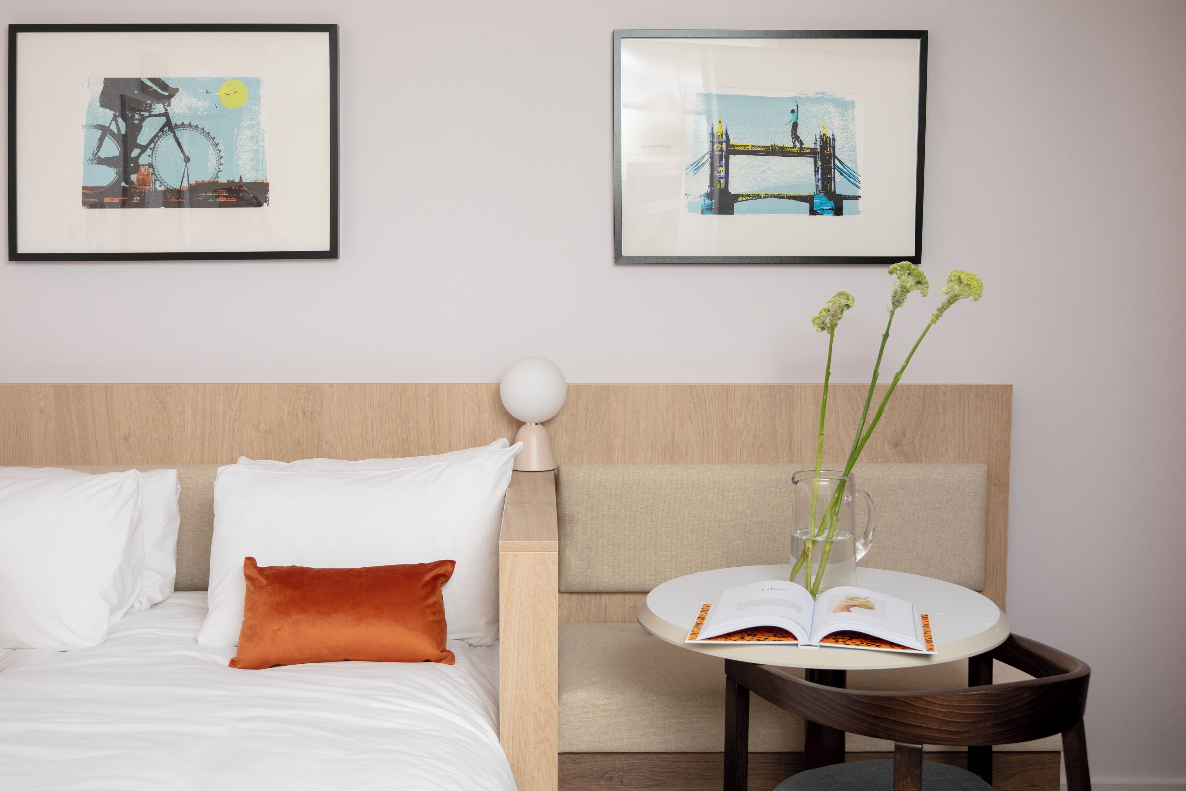 A hotel room with a bed, a round table with a book and a vase of flowers, and two framed pictures on the wall.