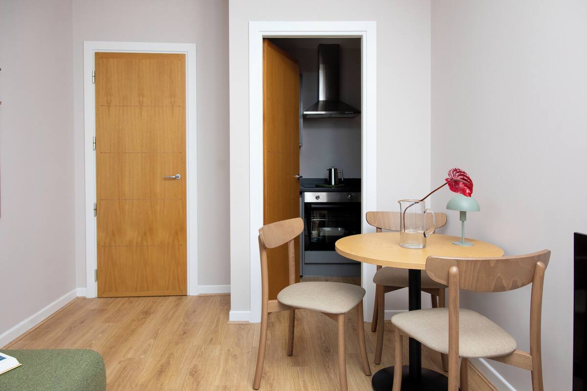 A small dining area with a round table, four chairs, a lamp, and a kitchen visible through an open door.