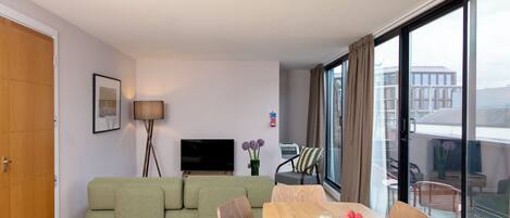A modern living room with a green sofa, a wooden dining table, and a flat-screen TV.