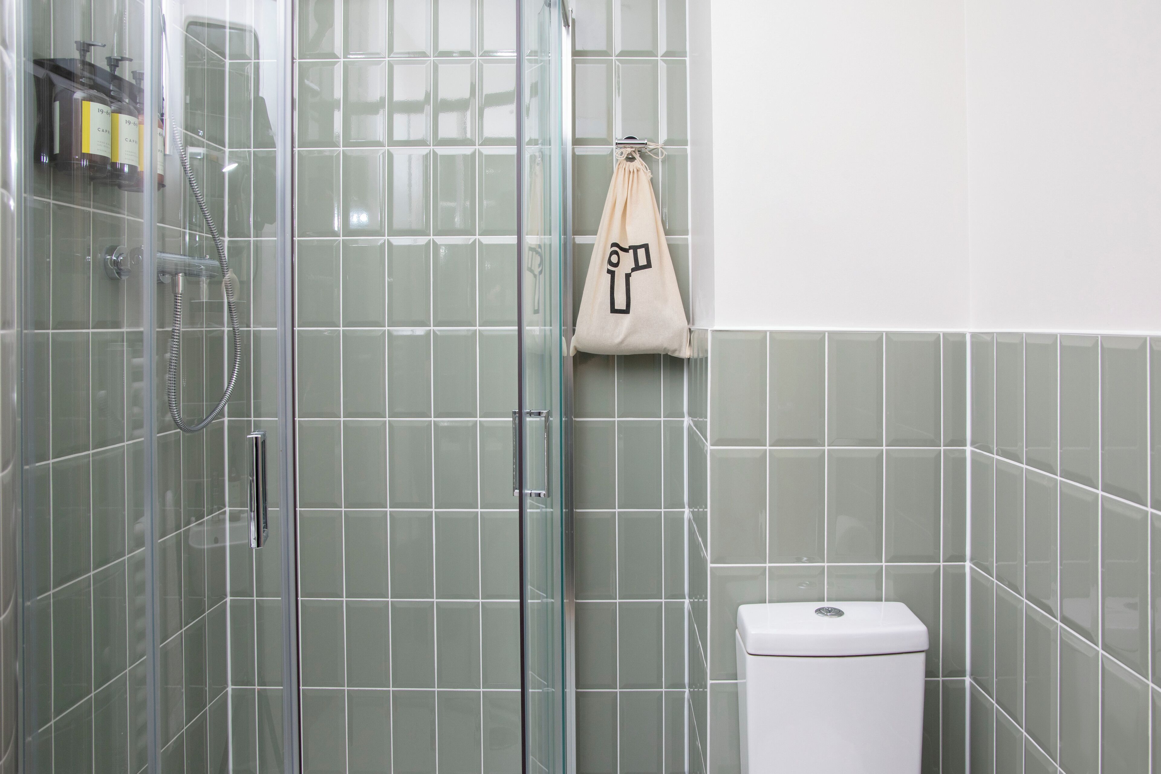 Une salle de bain avec une douche à parois de verre, des toilettes blanches et un sac beige accroché à un crochet.