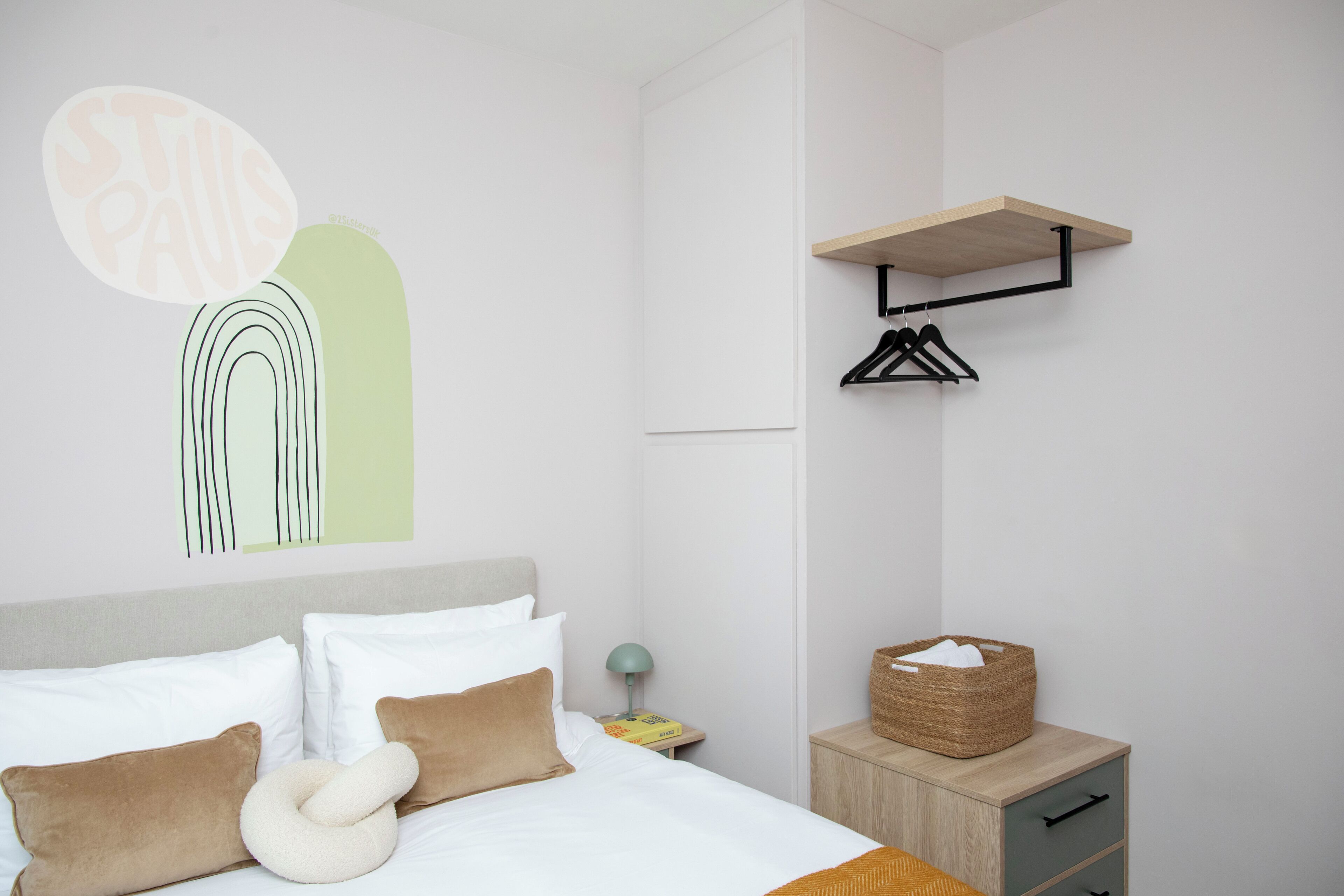 Une chambre d’hôtel moderne avec un lit soigneusement fait, une table de chevet avec une lampe, une petite table de nuit en bois et une étagère murale avec un panier.