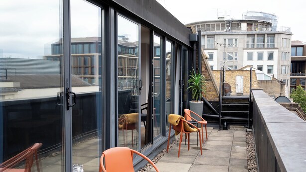 A modern outdoor seating area with glass doors, a potted plant, and a view of surrounding buildings. - Bob W London St. Paul's (London)