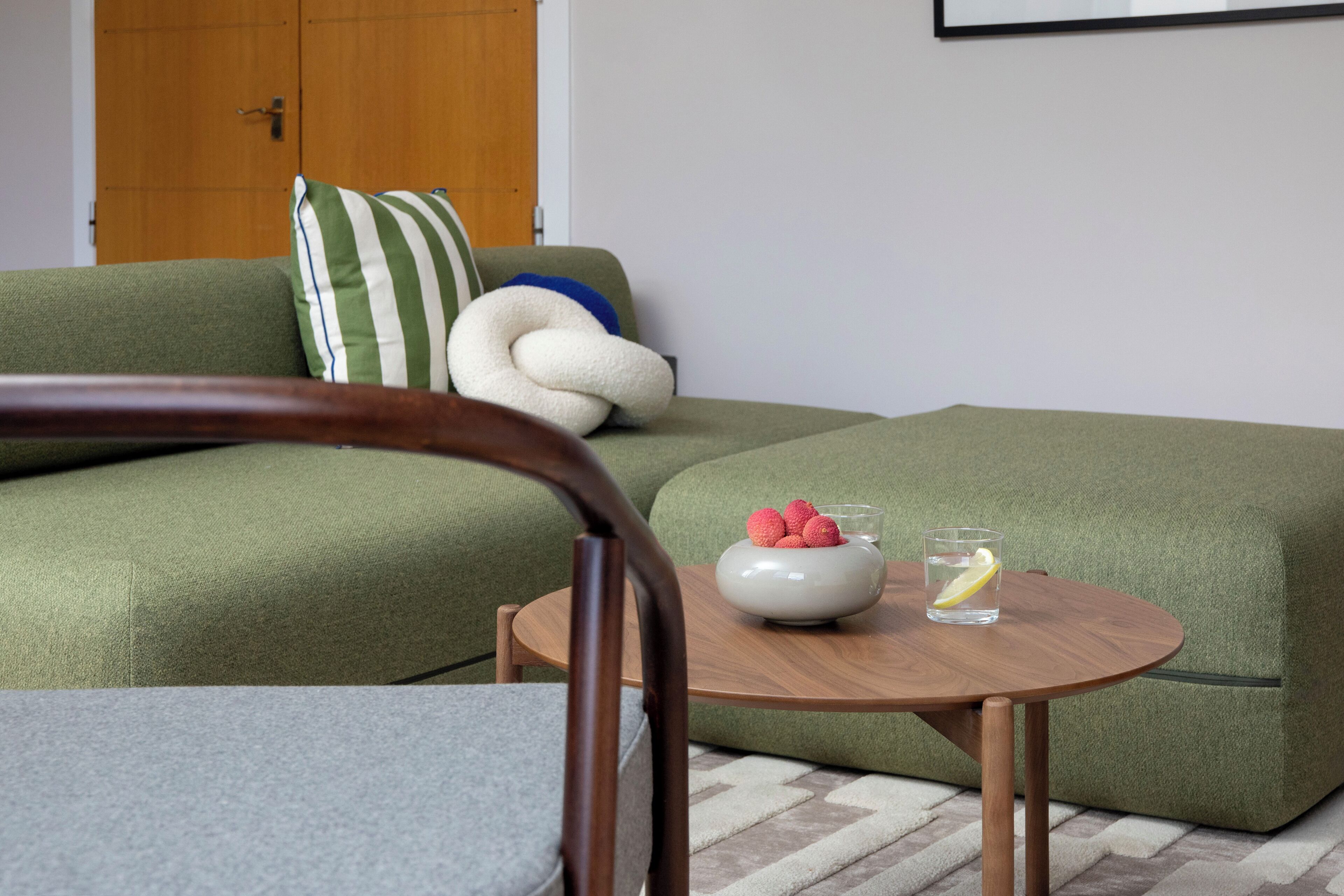 Ein grünes Sofa mit einem gestreiften Kissen, ein runder Holztisch mit einer Obstschale und einem Glas Wasser, und eine Holztür im Hintergrund.