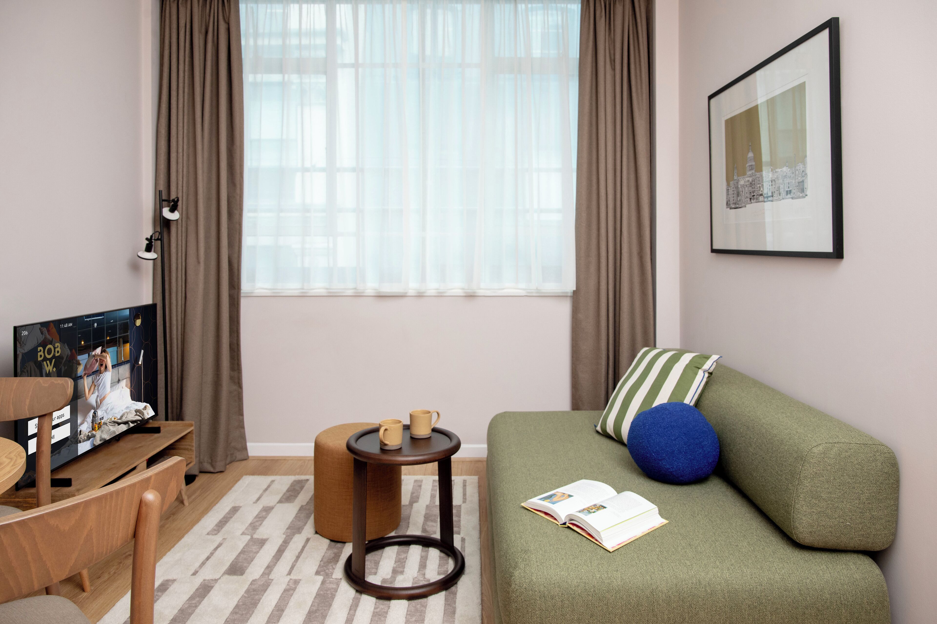 Een gezellige kamer met een groene bank, een klein rond tafeltje, een televisie en een raam met gordijnen.