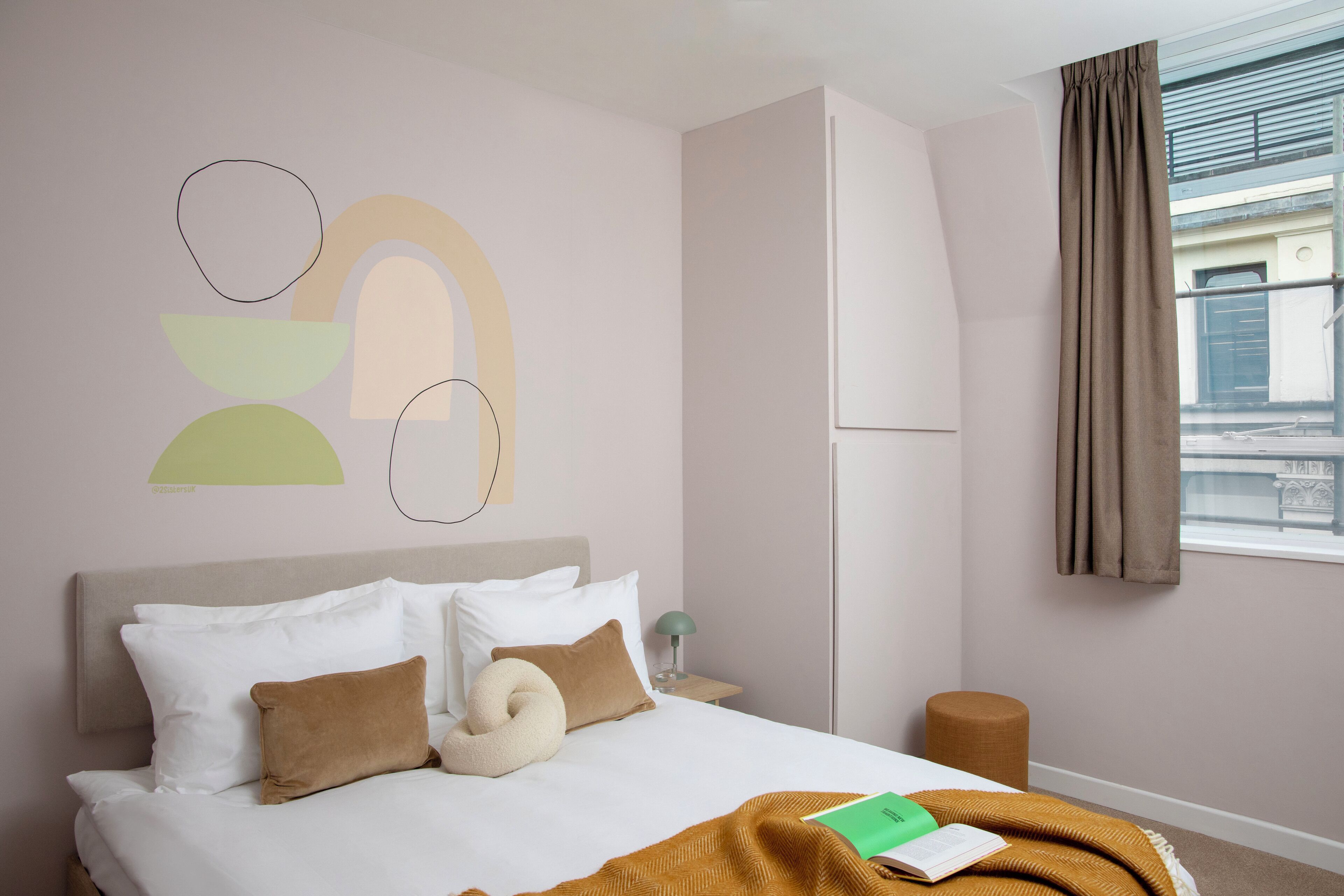 Hotelkamer met een bed, kussens, een deken, een boek, een lamp en een raam met gordijnen.