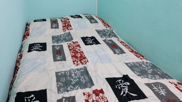 Une chambre avec un mur de couleur turquoise, un lit recouvert d’une courtepointe ornée de motifs japonais et un climatiseur fixé au mur.