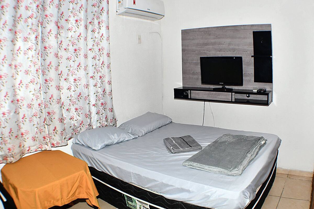 A bedroom with a bed, a TV mounted on the wall, a small table, and a curtain with a floral pattern.