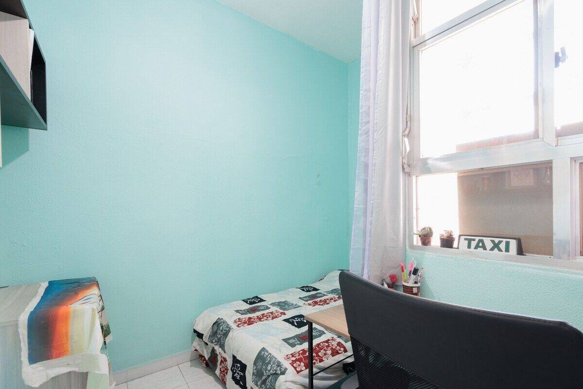 A small room with teal walls, a bed with a patterned bedspread, a desk with a computer, and a window with a 'TAXI' sign.