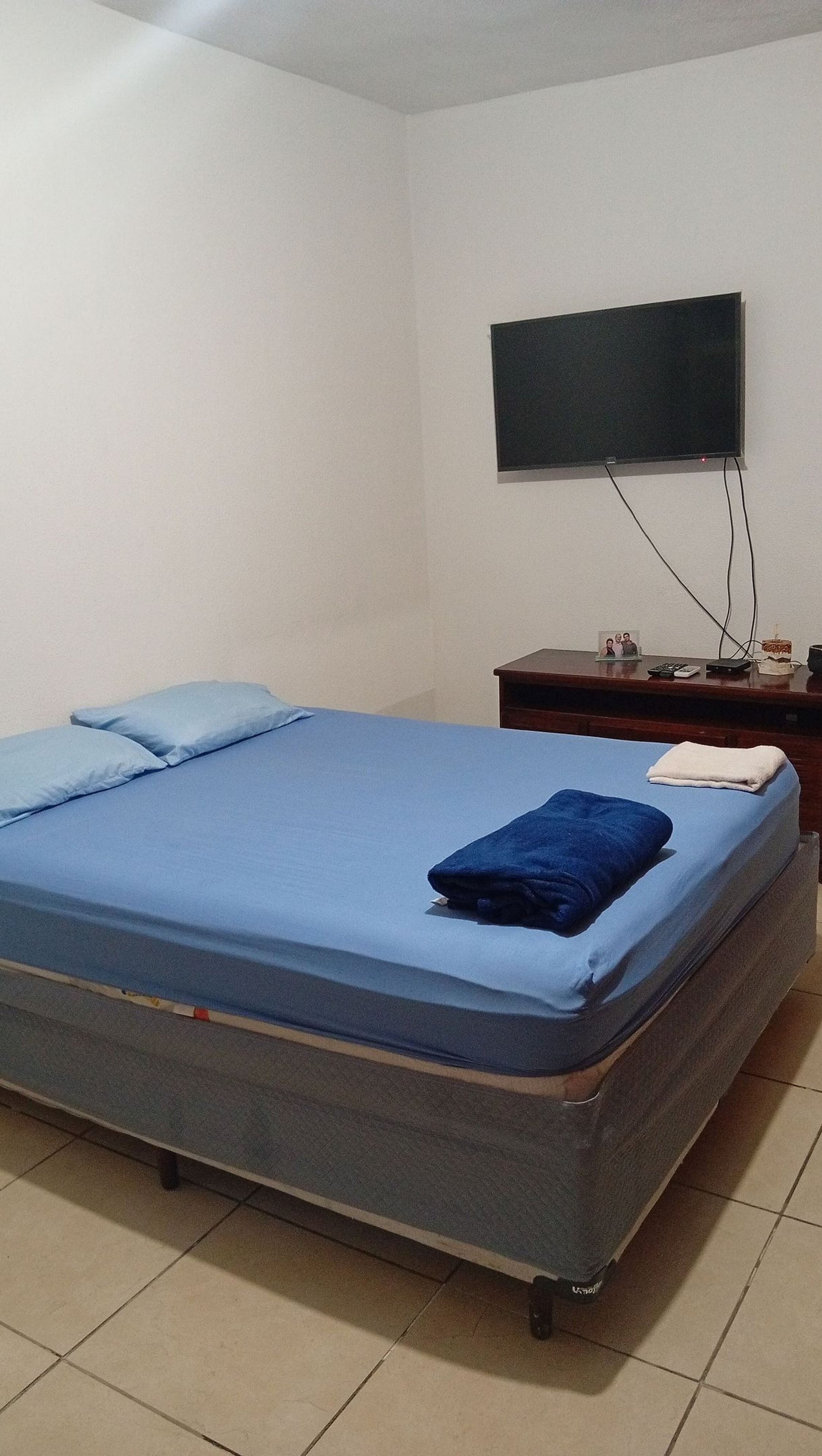 A bedroom with a single bed, a flat-screen TV mounted on the wall, a wooden nightstand with a lamp, and a small table with a picture frame and a clock.