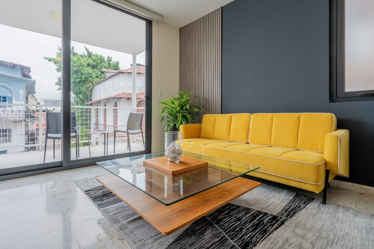 A modern living room with a yellow sofa, a glass coffee table, and a view of the city through large windows.