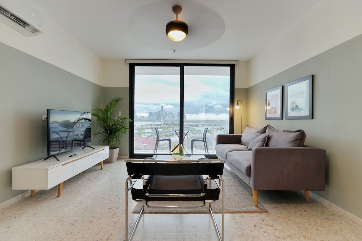 A modern living room with a large window, a black and white TV stand, a brown sofa, and a black table with a glass top.