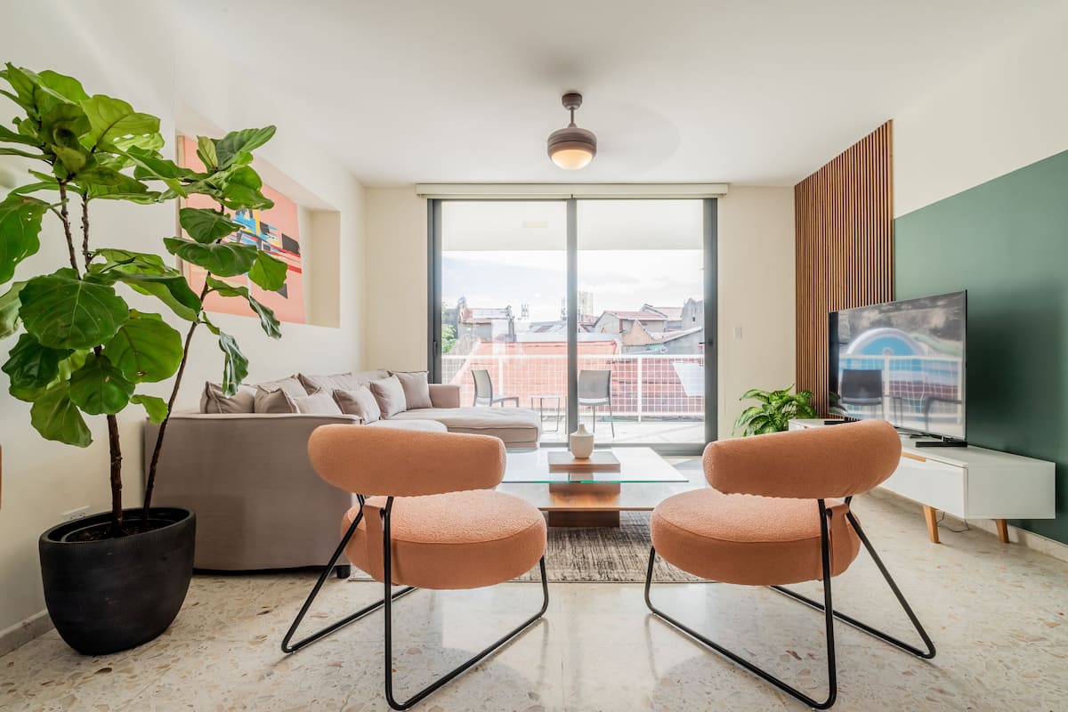 A modern living room with a sofa, two armchairs, a coffee table, a TV, and a large window with a city view.