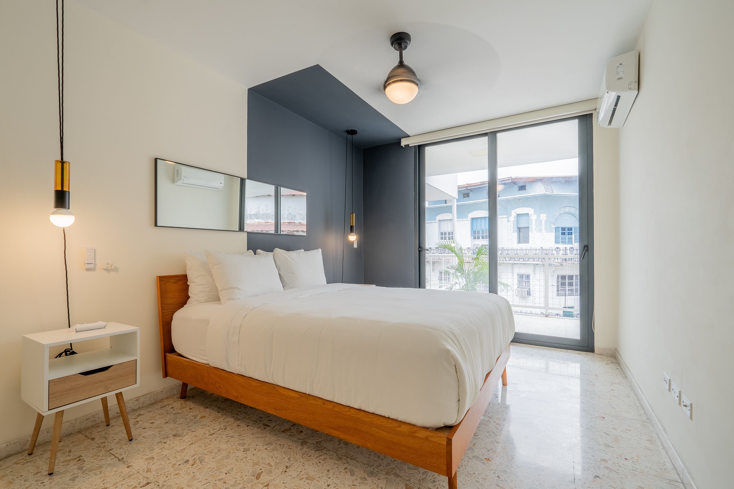 Une chambre à coucher avec un lit, une table de chevet, un miroir et une grande fenêtre donnant sur des immeubles.