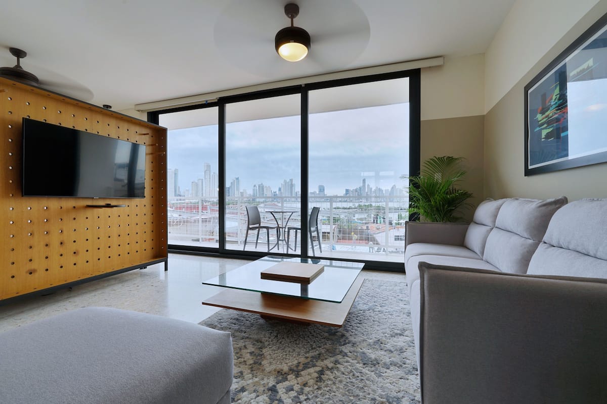 A modern living room with a large TV, a glass coffee table, and a sofa. There is a city view through large windows.