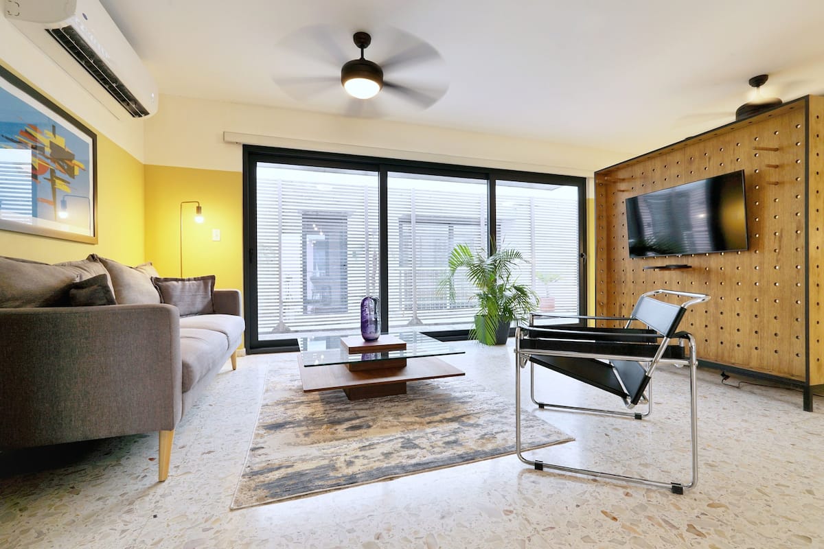 A modern living room with a sofa, a glass coffee table, a floor lamp, a TV mounted on a wooden wall, and a large window with blinds.