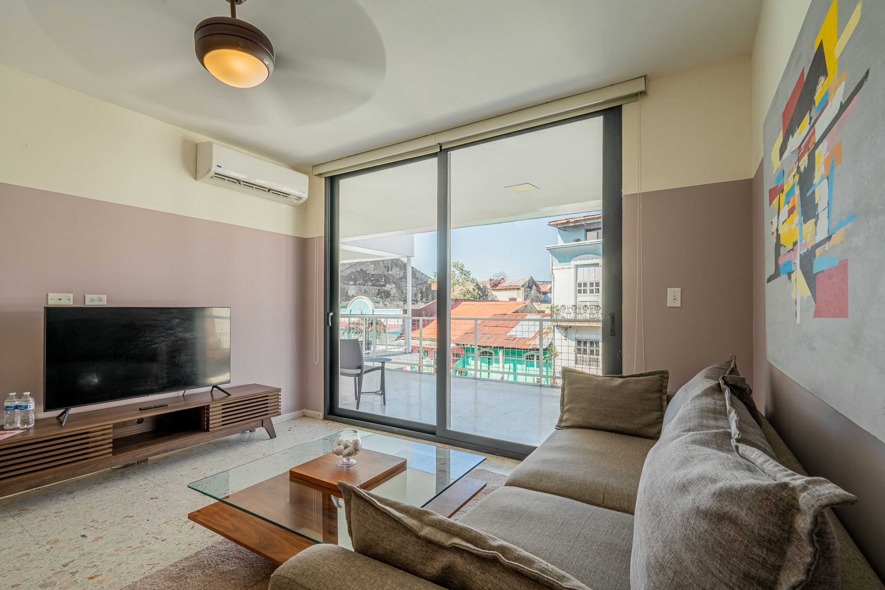 Una sala moderna con una televisión de pantalla plana, una mesa de centro de vidrio, un sofá y un amplio ventanal corredizo que da a un área exterior.
