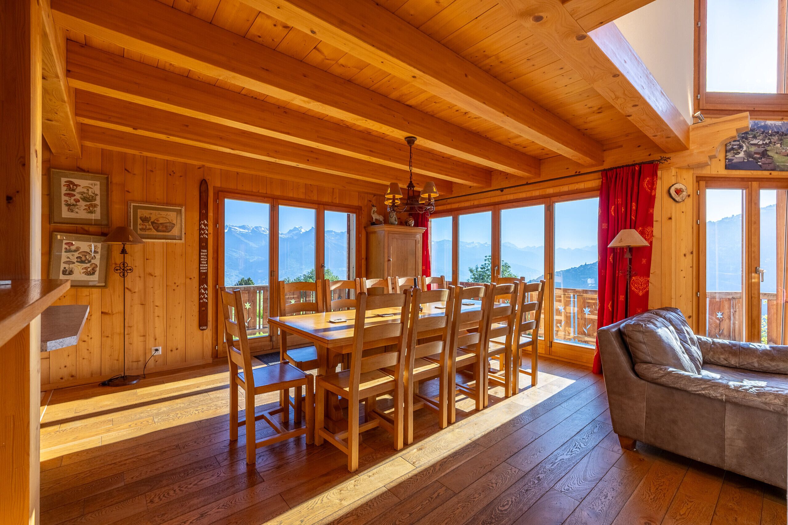A wooden interior with a dining table, chairs, a sofa, and large windows offering mountain views.