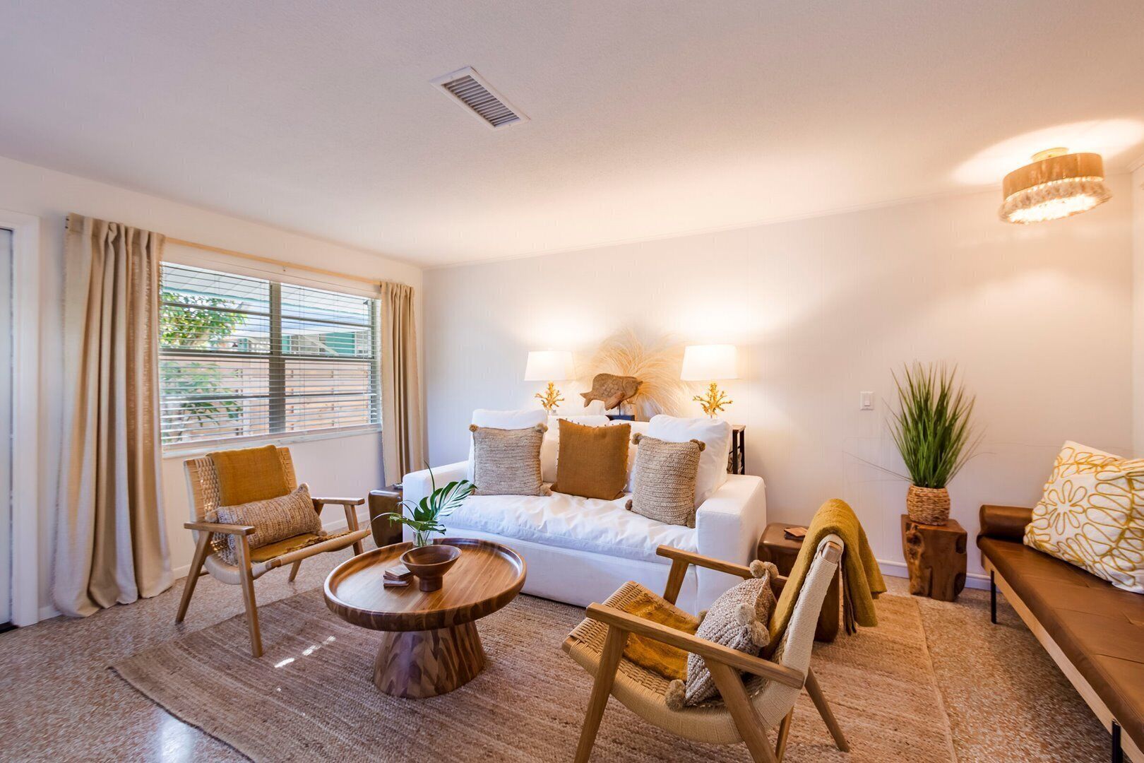 Ein gemütliches Wohnzimmer mit einem weißen Sofa, Holzstühlen, einem Couchtisch und einem großen Fenster mit Vorhängen.
