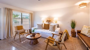 A cozy living room with a white sofa, wooden chairs, a coffee table, and a large window with curtains. - Barefoot Mermaid 4, steps to the beach (Anna Maria)