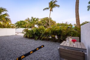 A patio area with a gravel ground, a wooden table with two wine glasses, a bottle of wine, and a vase with flowers. - Barefoot Mermaid 4, steps to the beach (Anna Maria)