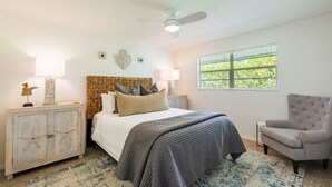 Ein Schlafzimmer mit Bett, Nachttischen, einem Sessel, Deckenventilator und einem Fenster mit Jalousien.