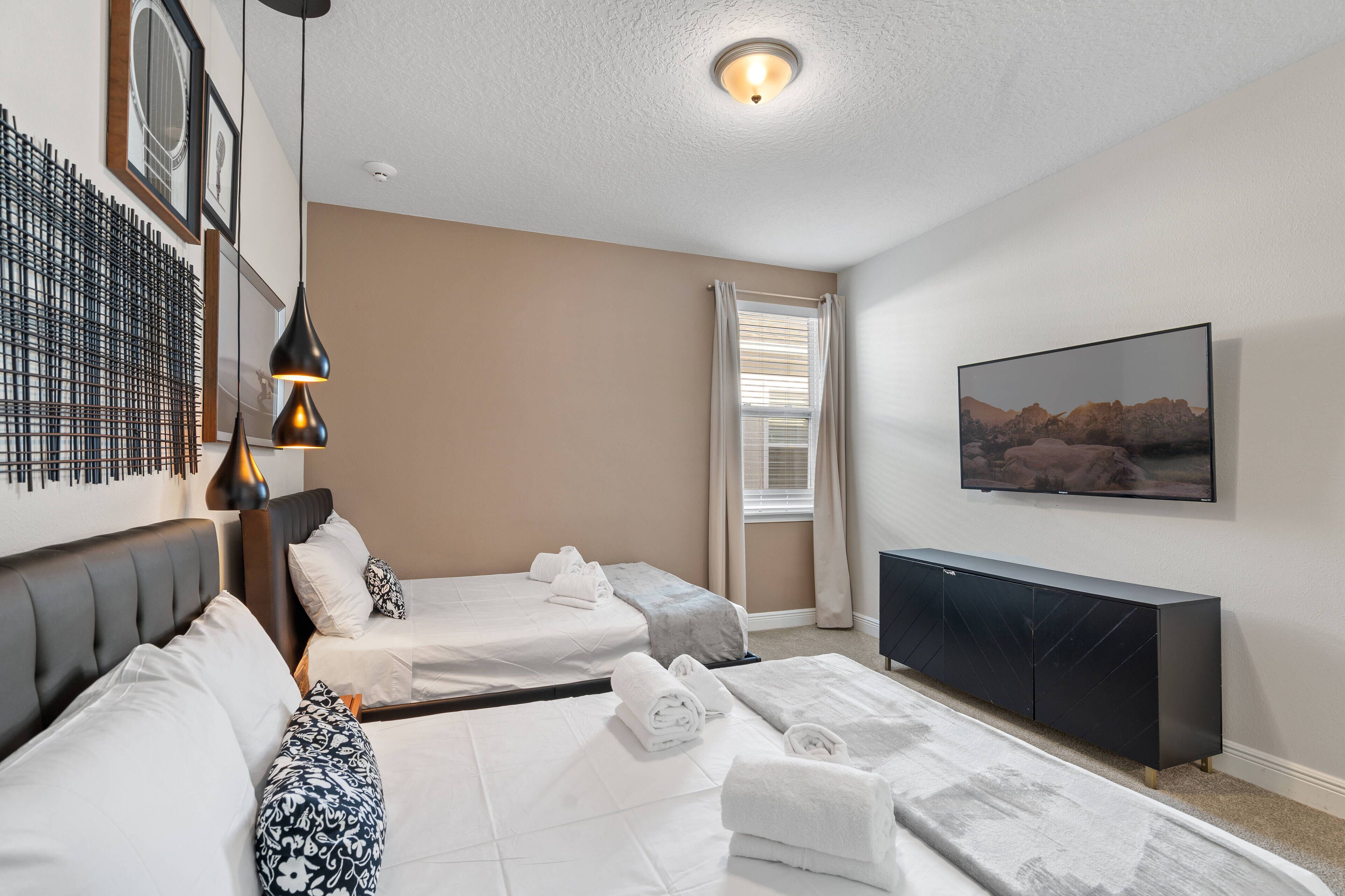 A hotel room with two beds, a black console table, a mounted TV, and a large window with blinds.