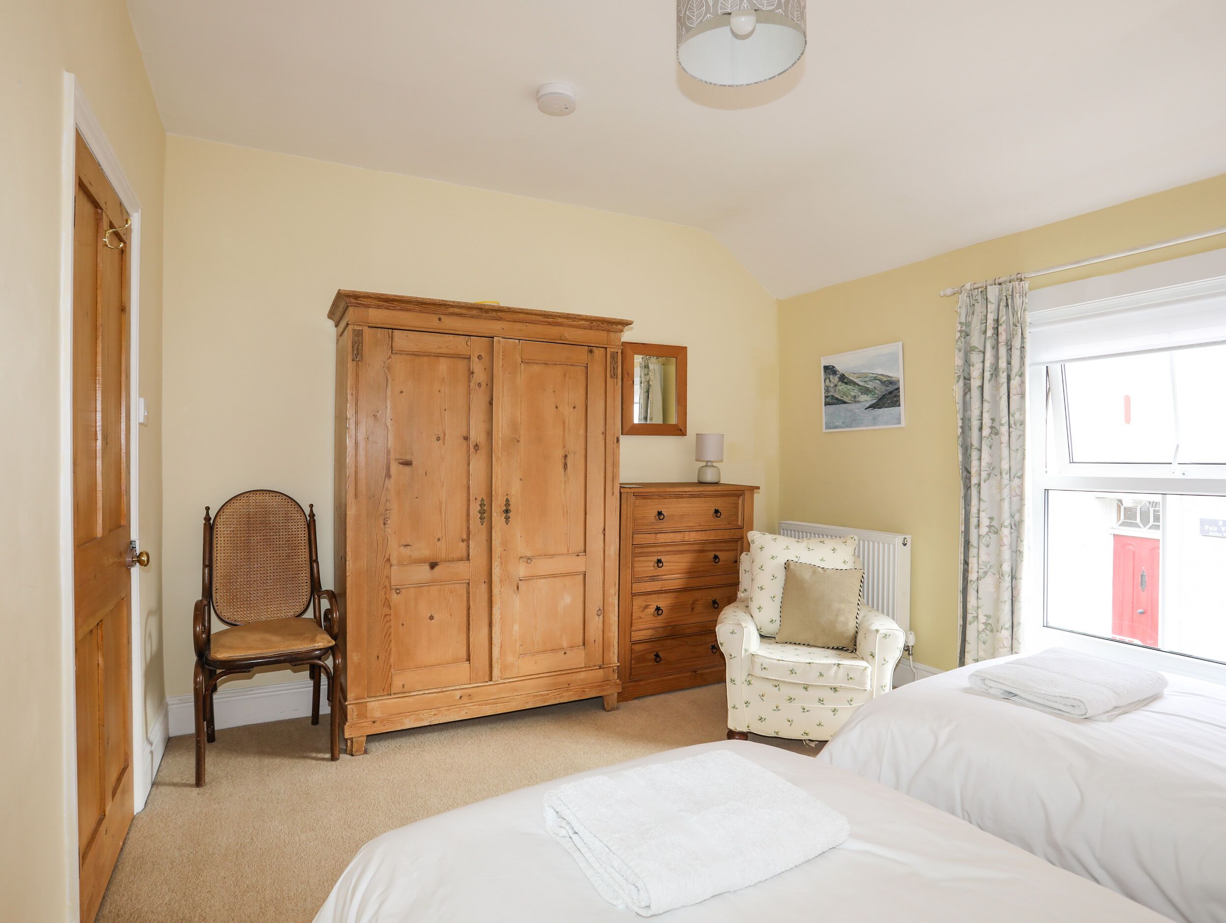 A bedroom with a wooden wardrobe, a chair, a dresser, and a bed with white linens.