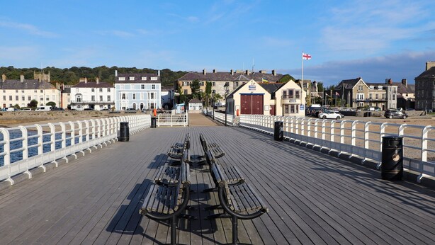 A wooden pier with benches, a white railing, and buildings in the background. - Amelie Cottage (Beaumaris)