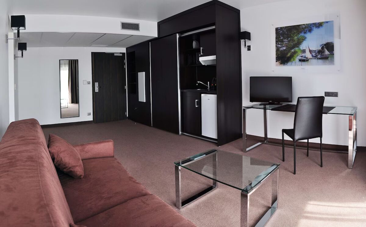 Een moderne hotelkamer met een bank, een glazen salontafel, een bureau met stoel, een televisie en een kledingkast.