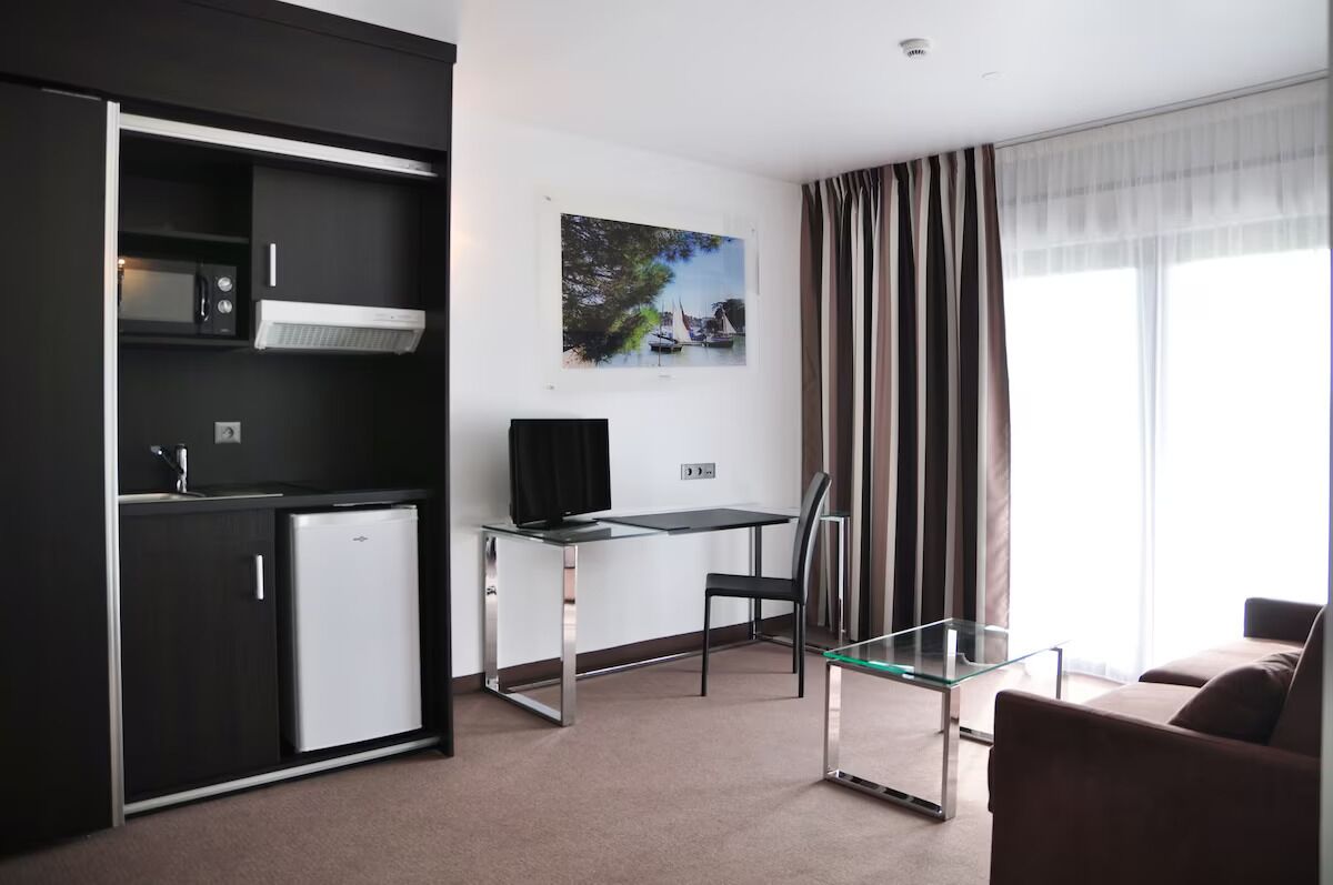 A modern hotel room with a kitchenette, a glass desk, a flat-screen TV, a glass coffee table, a brown sofa, and a large window with curtains.