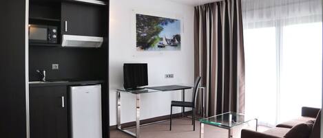 A modern hotel room with a kitchenette, a glass desk, a flat-screen TV, a glass coffee table, a brown sofa, and a large window with curtains.