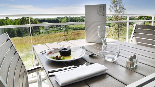 Mesa dispuesta con un plato de comida, cubiertos, una copa y un menú en un balcón con vistas a un paisaje.