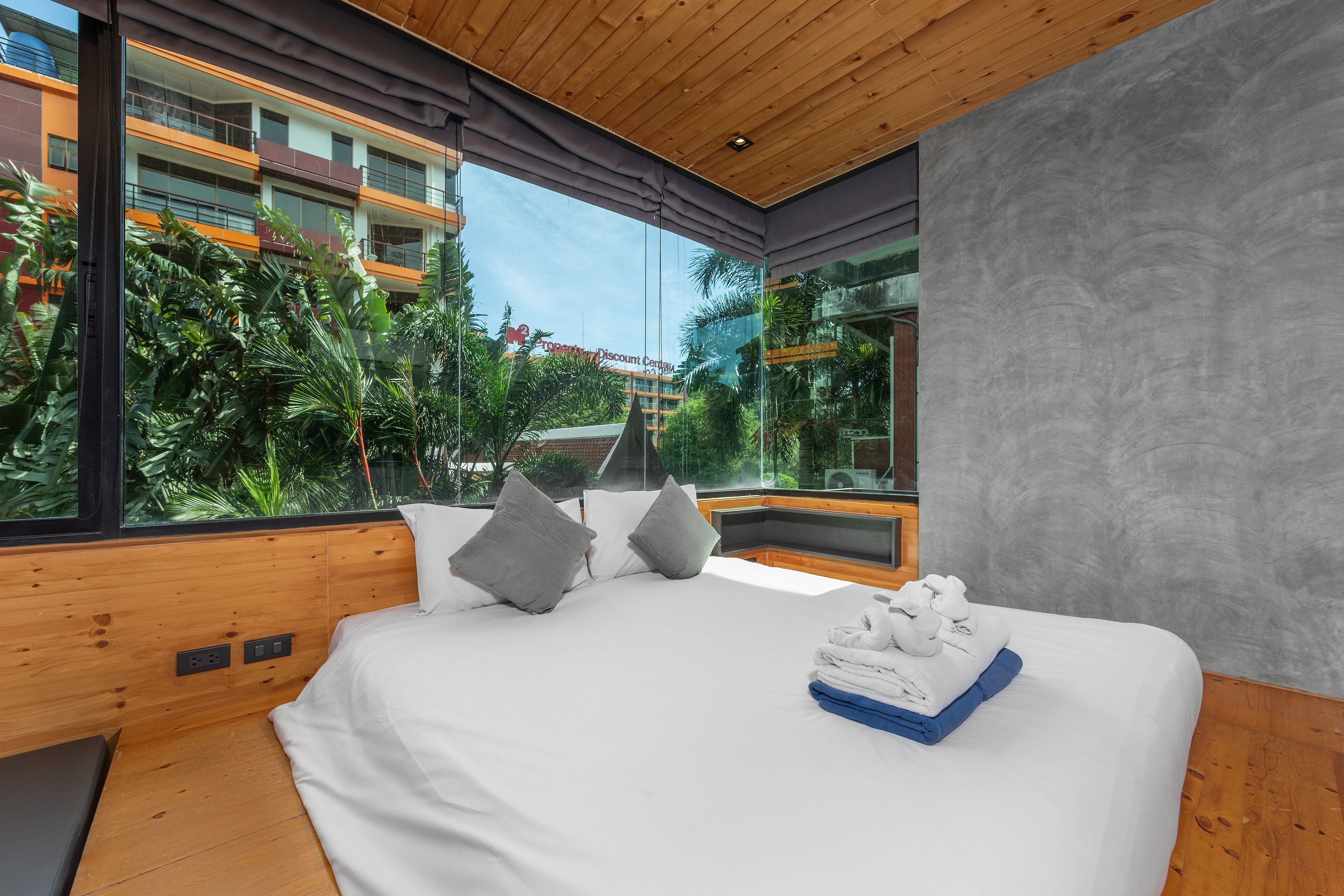 A modern hotel room with a large bed, wooden ceiling, and a view of a lush garden and building across the street.