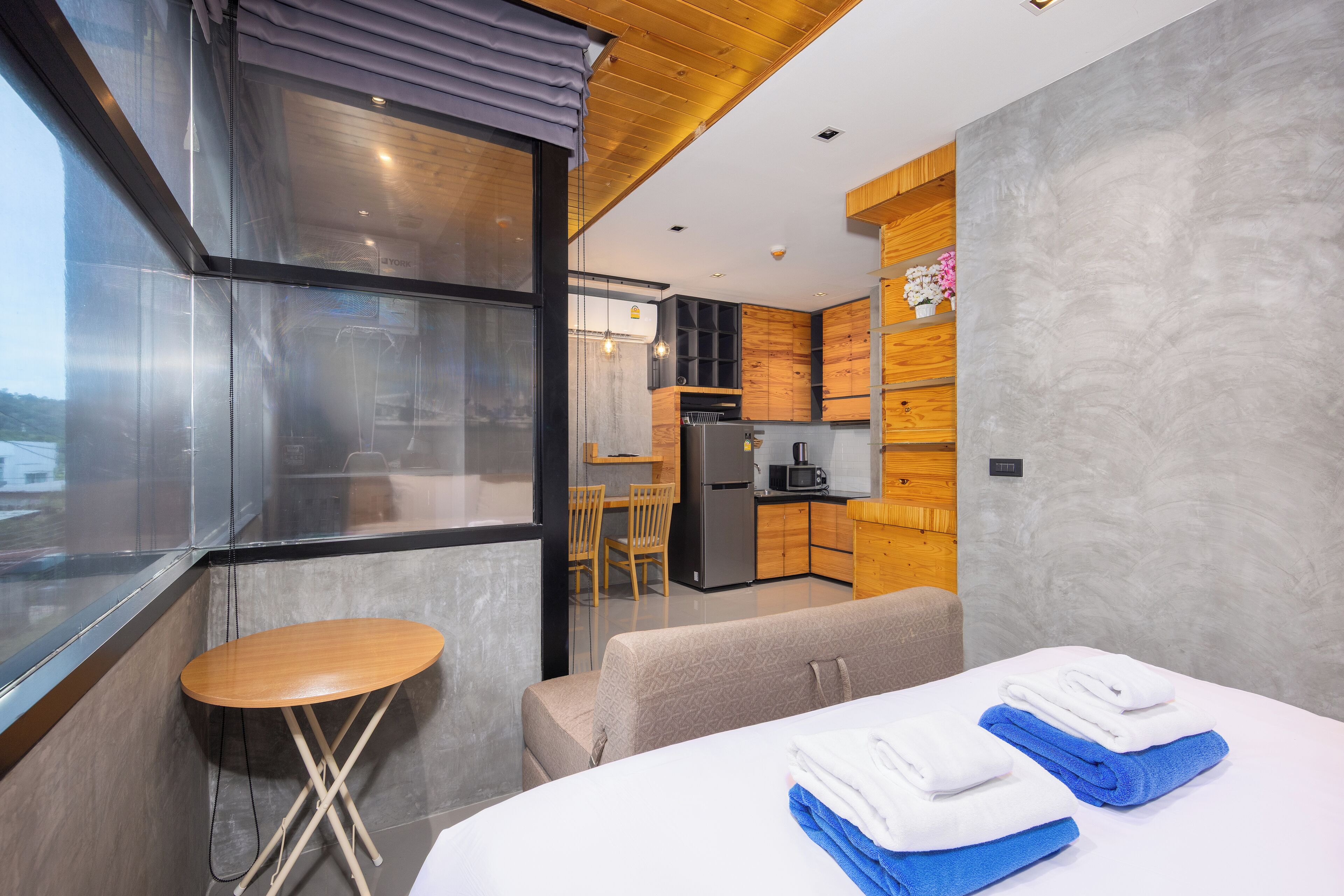 A modern hotel room with a large bed, a small wooden table, and a kitchenette in the background.