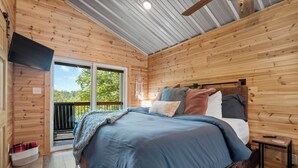A wooden cabin bedroom with a large bed, a small bedside table, and a view of the outdoors.