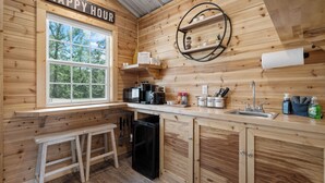 A wooden cabin-style kitchen with a 'Happy Hour' sign, a window, a sink, and a refrigerator.