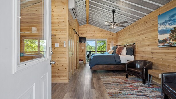 A wooden cabin room with a bed, a chair, a ceiling fan, and a large window.