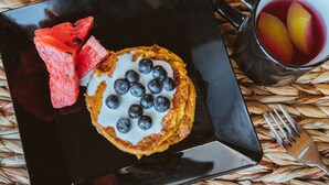 En svart, fyrkantig tallrik med ett bakverk toppat med blåbär, en skiva vattenmelon och en glasmugg med fruktskivor.