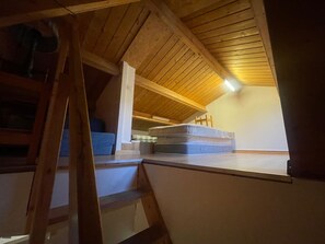 Une chambre aménagée dans les combles, avec un plafond en pente, des poutres en bois, un matelas posé sur le sol et une échelle.