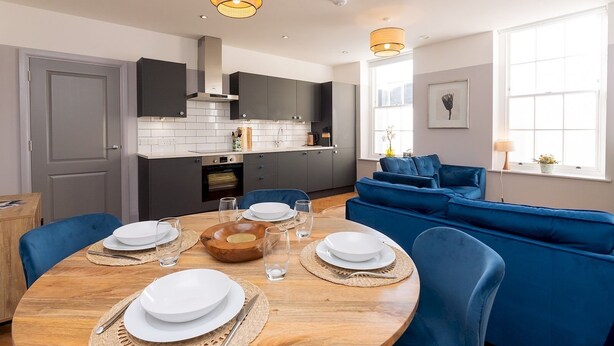 A modern kitchen with grey cabinets, a built-in oven, and a dining area with a round wooden table set for four. - Stunning 2 bed retreat in the heart of Bath - Pass the Keys (Bath)
