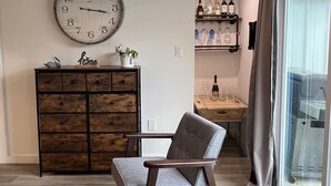 A modern living room with a large wall clock, a wooden cabinet, a grey sofa, and a dining area with wine bottles and glasses.