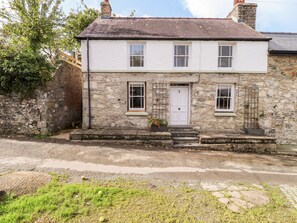 A two-story stone house with a white door and windows, a small porch, and a stone wall to the left.