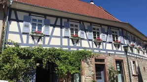 A traditional half-timbered building with white shutters and red flowers in window boxes, stone walls, and a clear blue sky. - SEVORAGE (Walzbachtal)