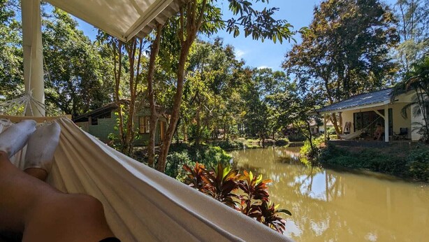Un hamac installé sous un espace ombragé, avec vue sur une étendue d’eau calme, entourée de verdure et d’un petit bâtiment.