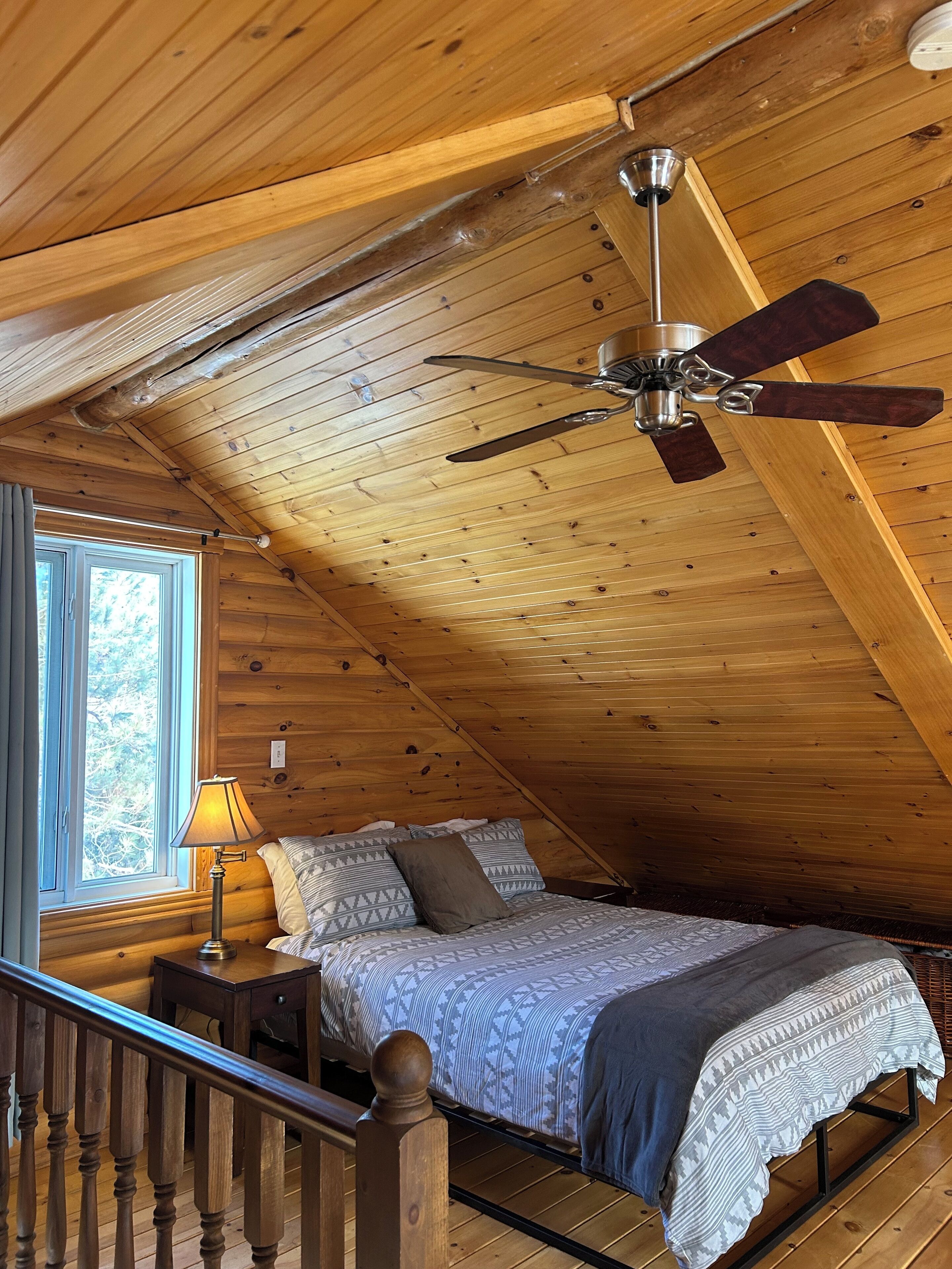 Un dormitorio acogedor en el ático con techo de madera, una cama con ropa de cama estampada, una mesita de noche y un ventilador de techo.