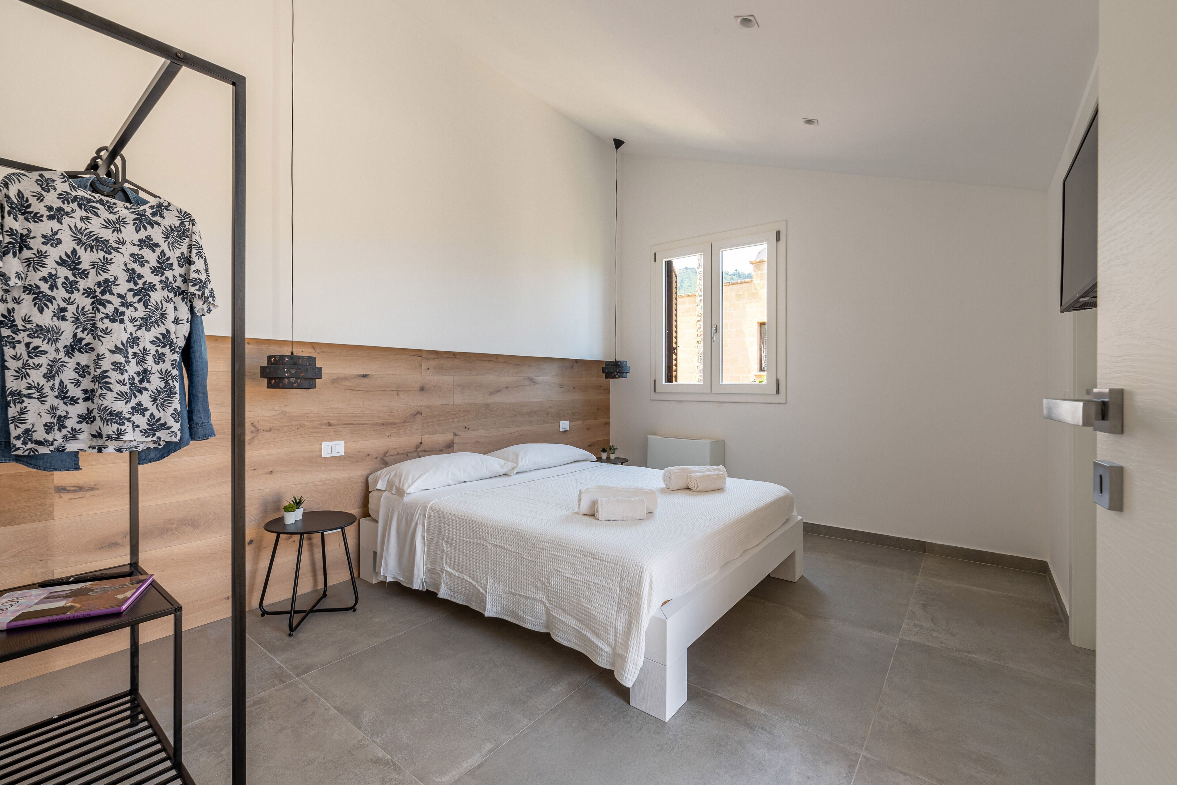 A modern bedroom with a wooden headboard, a bed with white linens, a small bedside table with a lamp, and a window providing natural light.