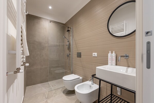 Un baño moderno con ducha de vidrio, inodoro blanco, bidé y lavamanos con artículos de aseo.